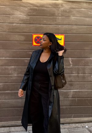 Mujer con abrigo y pantalones de cuero negro de pie frente a una puerta de garaje cerrada con una señal amarilla y roja de prohibido estacionar detrás de ella.