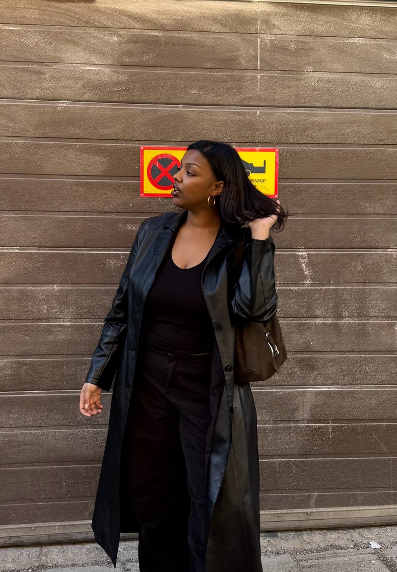 Femme en manteau et pantalon en cuir noir debout devant une porte de garage fermée avec un panneau jaune et rouge d'interdiction de stationnement derrière elle.