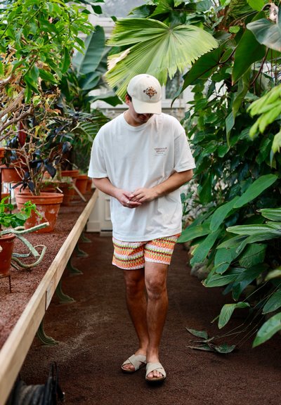 Joven con gorra blanca, camiseta blanca de gran tamaño, pantalones cortos coloridos y sandalias, caminando por un invernadero con grandes plantas tropicales y vegetación en macetas.