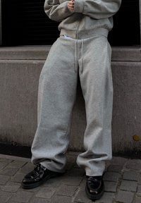 Personne portant un pantalon de survêtement gris surdimensionné et un sweat à capuche assorti zippé, debout sur un trottoir pavé avec des chaussures en cuir noir.