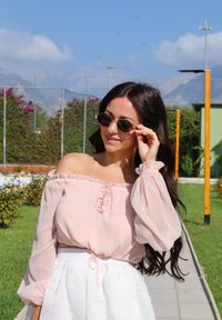 Blusa rosa clara, com decote off-the-shoulder, com babados e mangas compridas, combinada com uma saia branca de cintura alta. Os acessórios incluem óculos de sol escuros.