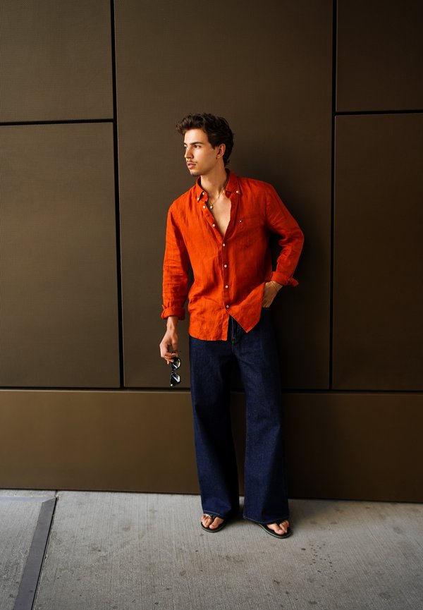 Mand i en opknappet orange skjorte, mørke vide jeans og sandaler står op ad en brun væg, holder solbriller og ser sidelæns.