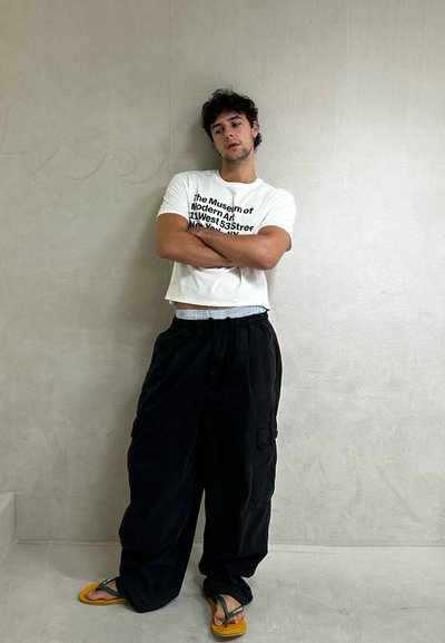 Camiseta blanca recortada con texto negro, pantalones cargo negros, chanclas amarillas, de pie contra una pared gris clara. Atuendo informal con un ajuste relajado.