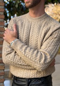 Homme portant un pull beige en maille torsadée et une montre-bracelet, les bras croisés, debout à côté d'un mur de briques avec un arrière-plan extérieur flou.