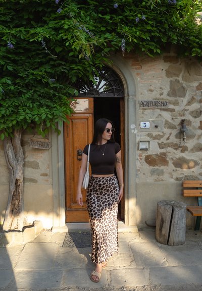 Top corto marrón, falda larga de estampado de leopardo, gafas de sol, de pie en una entrada de piedra rústica bajo la vegetación. Puerta de madera con un cartel cercano.