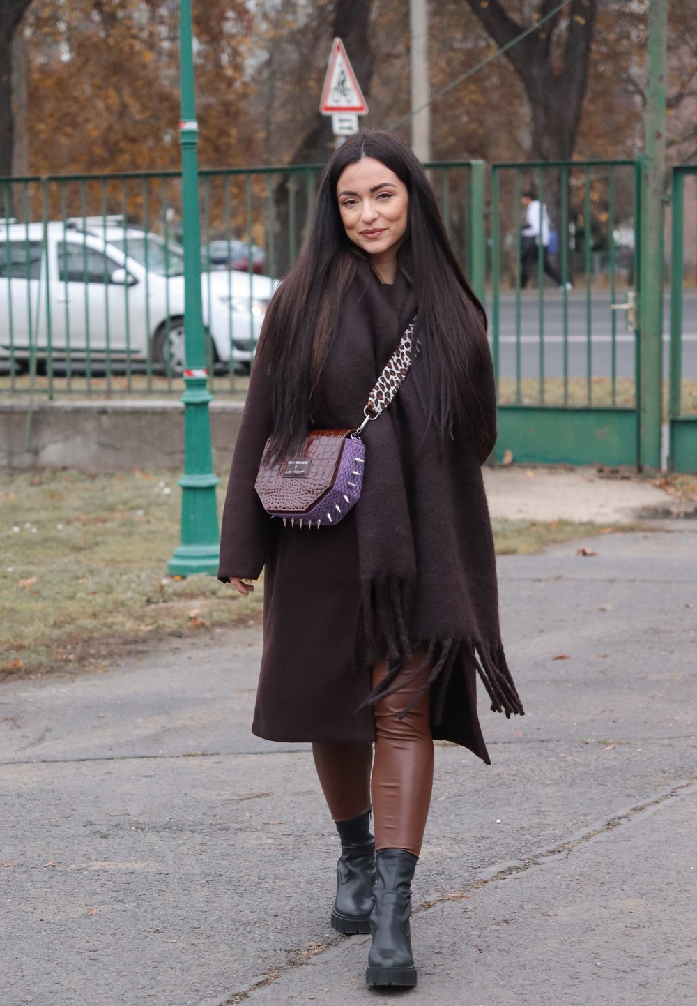 Rjavi pleteni plašč z resicami, rjave legice iz umetnega usnja, črni robustni čevlji in vijolična torbica z vzorcem in srebrnimi nitkami.