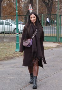 Rjavi pleteni plašč z resicami, rjave legice iz umetnega usnja, črni robustni čevlji in vijolična torbica z vzorcem in srebrnimi nitkami.