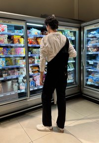 Persona con un overol negro y una camisa de manga larga blanca, mirando las vitrinas de alimentos congelados con empaques coloridos en un pasillo de supermercado.