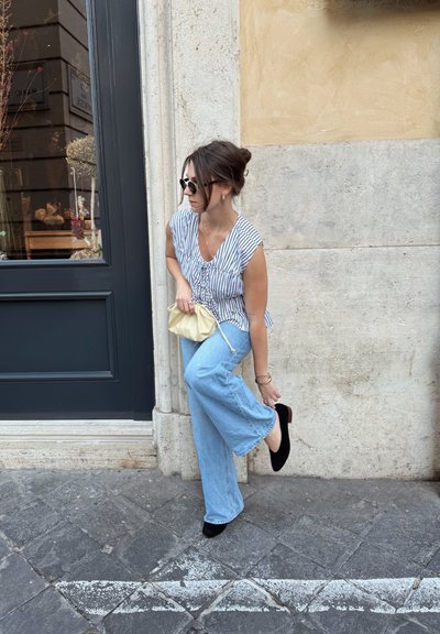 Blusa de rayas de manga corta; jeans de pierna ancha de color azul claro; zapatos negros; sosteniendo un pequeño bolso dorado; posando cerca de una pared de piedra.