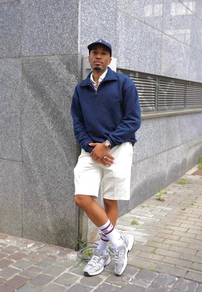 Hombre con gorra azul marino, jersey azul marino, pantalones cortos blancos y zapatillas apoyado contra una pared de piedra gris en una acera de la ciudad.