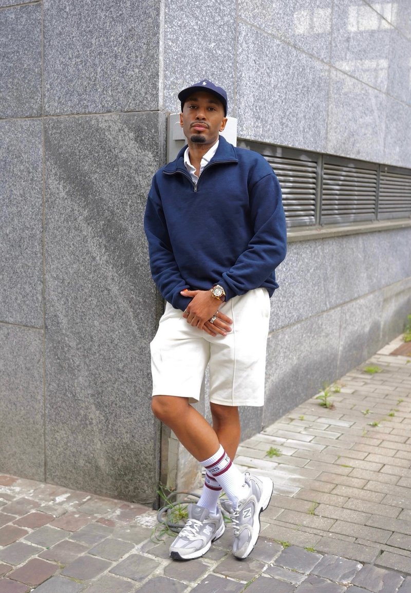Mand i navy-kasket, navy-pullover, hvide shorts og sneakers læner sig op ad en grå stenmurst på en byfortov.