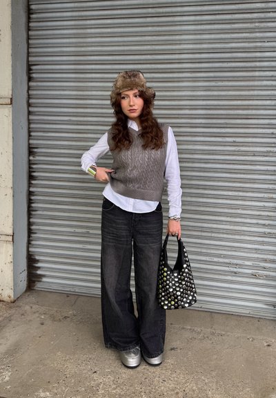 Chaleco gris de punto con un botón blanco, jeans anchos negros y zapatos de plataforma plateados brillantes. Bolsa negra con lunares, el fondo es metálico.