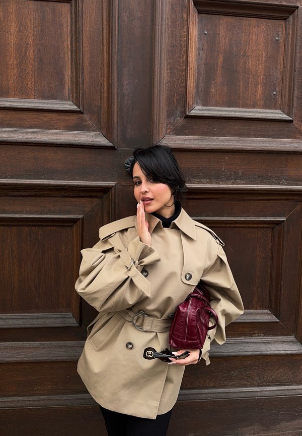 Una mujer con una gabardina beige con cinturón posa delante de una puerta de madera oscura, sosteniendo un bolso burdeos y unas gafas de sol.