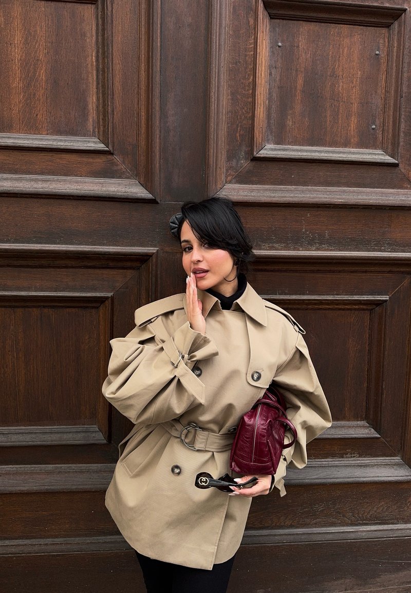 Une femme en trench-coat beige ceinturé pose devant une porte en bois sombre, tenant un sac à main bordeaux et des lunettes de soleil.