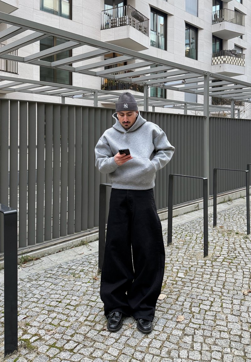 Jeune homme en sweat à capuche gris et pantalon large noir se tient sur un chemin pavé, regardant son téléphone près d'un bâtiment moderne avec une rambarde métallique et des balcons.