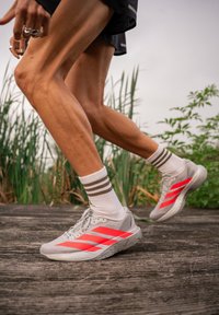 Gambe di un corridore a metà passo, con calze bianche a strisce marroni e scarpe da ginnastica grigie con strisce rosse brillanti, su un sentiero di legno all'aperto.