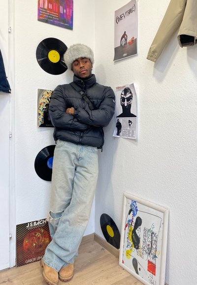 Chaqueta de plumas negra, jeans desgastados color azul claro, botas marrones y gorro de piel gris. La decoración de la pared incluye discos de vinilo y obras de arte coloridas.