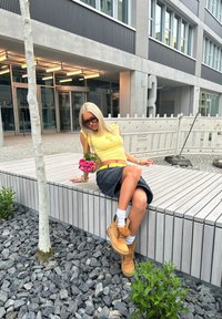 Nainen istuu puupenkillä keltapaitaisena ja mustassa hameessa, pitäen pinkkejä ruusuja käsissään. Hänellä on päällään ruskeat saappaat ja valkoiset sukat, moderni rakennus taustanaan.