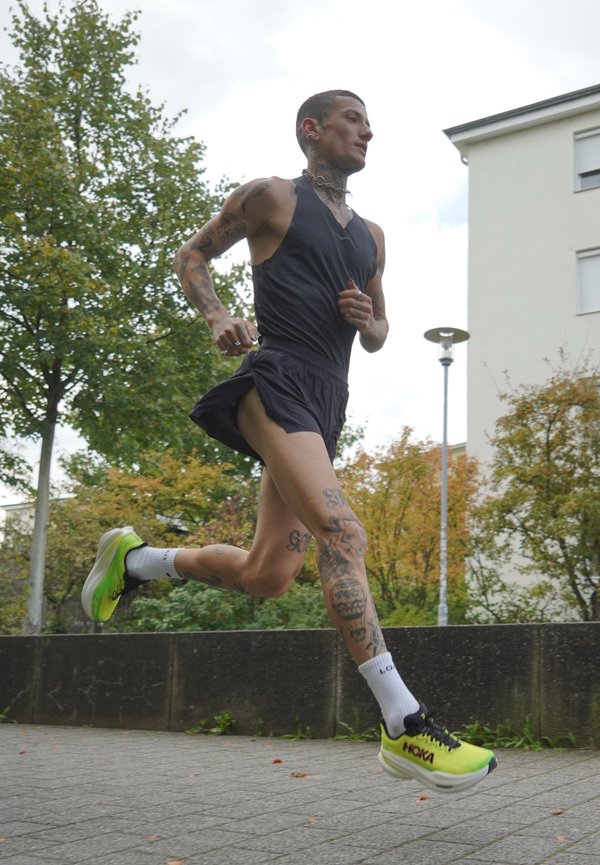 Løper med tatoveringer iført svart treningsdrakt og neongule Hoka-sneakers som jogger ute på en asfaltsti nær trær og bygninger.