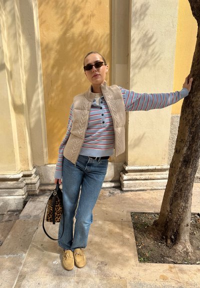 Mujer con gafas de sol, suéter de rayas, chaleco beige, vaqueros azules y zapatos marrones, apoyada en un árbol con una mano y sosteniendo un bolso con estampado de leopardo.