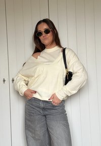 Prevelik pulover kremaste barve z visokim ovratnikom in izrezanimi detajli, kombiniran s sivimi širokimi kavbojkami in majhno črno torbico.