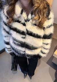 Femme portant une veste rayée noire et blanche en peluche, un pantalon noir et des chaussures noires pointues, tenant un sac à main noir à l'intérieur.