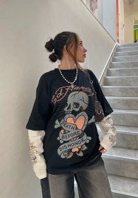 Svart oversized t-skjorte med hodeskalle- og hjertegrfikk, hvite langermede skjorter, grå jeans og en liten svart veske. Perlekjede som aksent.