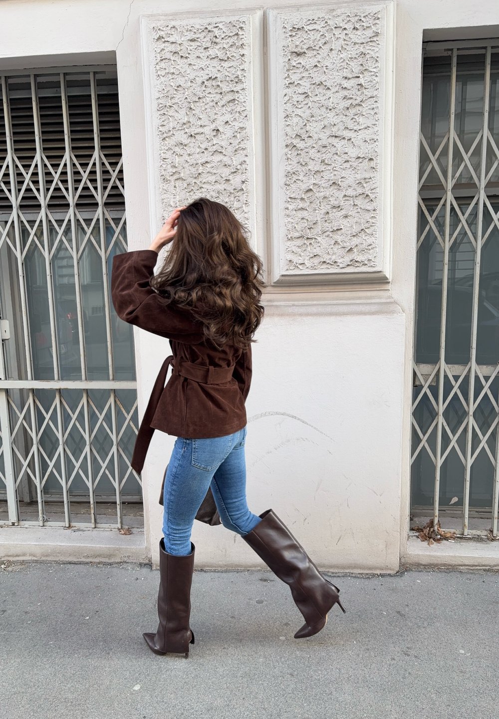 Femme aux longs cheveux ondulés, vêtue d'une veste marron, d'un jean bleu et de hautes bottes marron, marchant devant un mur blanc texturé avec des fenêtres grillagées.