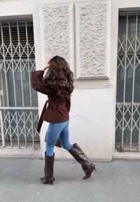 Femme aux longs cheveux ondulés, vêtue d'une veste marron, d'un jean bleu et de hautes bottes marron, marchant devant un mur blanc texturé avec des fenêtres grillagées.