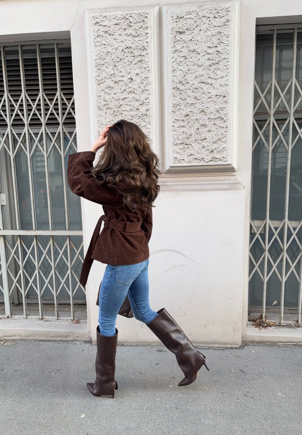 Hosszú, hullámos hajú nő barna dzsekiben, kék farmerben és magas barna csizmában, aki egy rácsos ablakokkal ellátott, texturált fehér fal mellett sétál el.