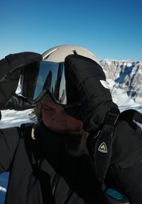 Des gants noirs ajustent des lunettes de ski miroitées au-dessus d'un casque beige. Une veste sombre contraste avec un ciel bleu et des montagnes enneigées en arrière-plan.