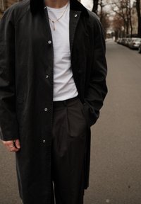 Manteau long noir avec col, boutons-pression, porté sur une chemise blanche et un pantalon gris. Bague en argent et collier chaîne visibles.