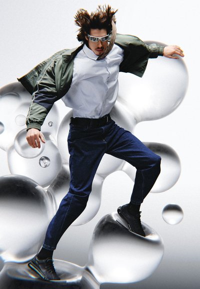 Figura masculina vestida con una chaqueta verde y negra sobre una camisa blanca y jeans oscuros; fondo gris.