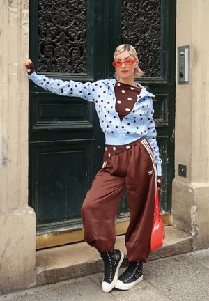 Persona apoyada contra una puerta oscura y ornamentada, vestida con chaqueta azul de lunares, camiseta marrón de lunares, pantalones marrones, zapatillas negras, gafas de sol rojas y sosteniendo una bolsa roja.