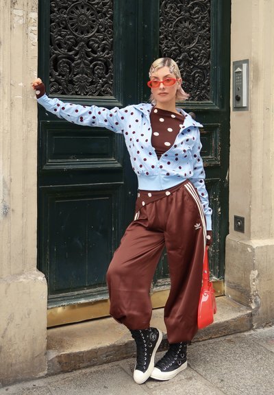 Persona apoyada contra una puerta oscura y ornamentada, vestida con chaqueta azul de lunares, camiseta marrón de lunares, pantalones marrones, zapatillas negras, gafas de sol rojas y sosteniendo una bolsa roja.