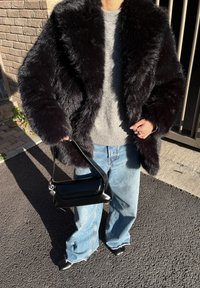 Personne portant un manteau en fausse fourrure noire, un pull gris, un jean bleu ample, des chaussures noires, et tenant un sac à main en cuir noir à l'extérieur sous le soleil.