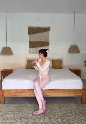 Mujer con flor en el cabello sentada en una cama de madera sosteniendo un vaso, en una habitación con un tapiz de pared tejido y dos lámparas colgantes.