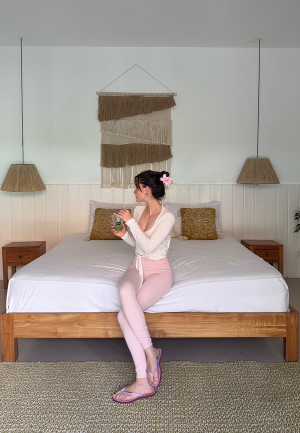 Frau mit Blume im Haar, die auf einem Holzbett sitzt und ein Glas hält, in einem Zimmer mit gewebtem Wandbehang und zwei Hängeleuchten.