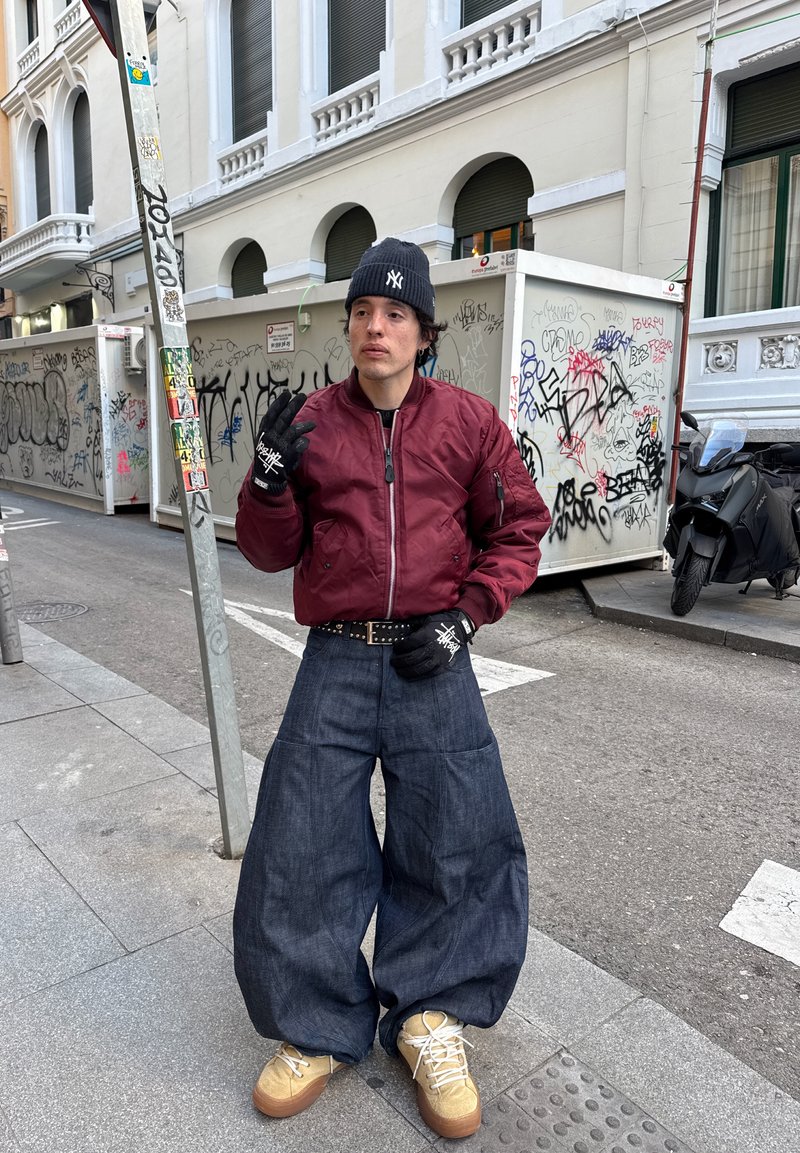 Homme en veste bordeaux, jean large foncé, gants noirs et bonnet bleu marine NY, debout dans une rue urbaine avec des graffitis et un scooter en arrière-plan.