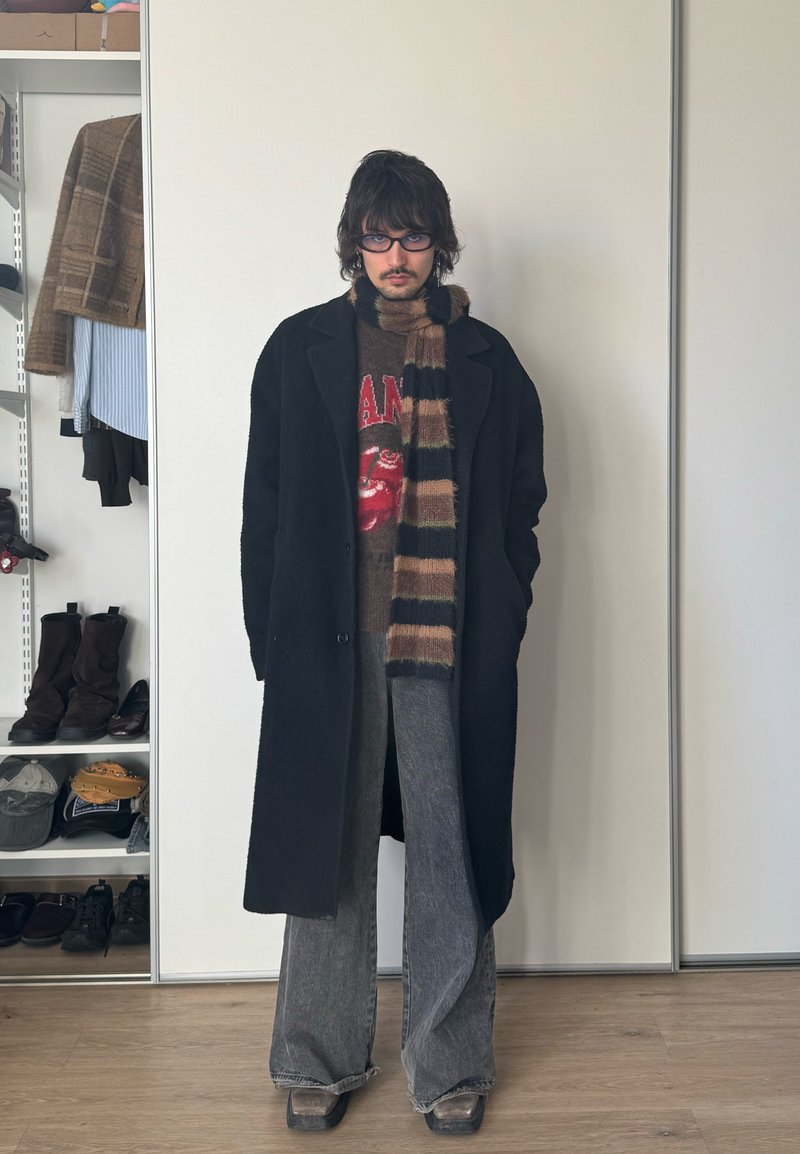 Inimene, kes kannab musta mantlit, triibulist salli, graafilist kampsunit, lahtisi halli pükse ja prille, seisab toas riidekapi lähedal, kus on riided ja kingad.