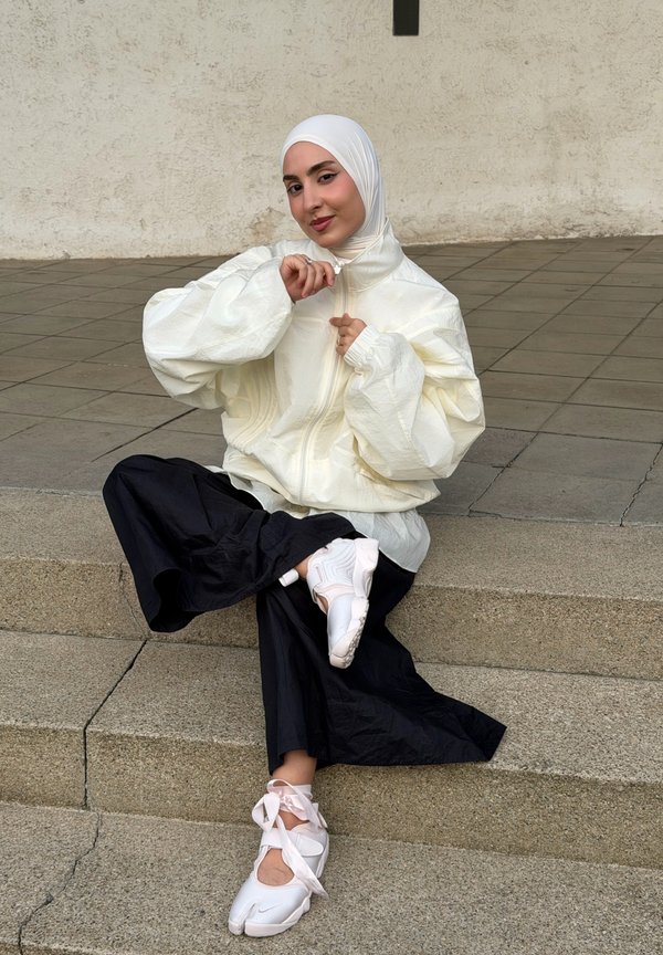 Jeune femme portant un hijab blanc, une veste blanche, un pantalon large noir et des baskets blanches, assise sur des marches en pierre en plein air.