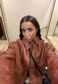 Jeune femme aux longs cheveux foncés portant une veste en cuir marron et un sac bandoulière noir prenant un selfie à l'intérieur près d'un escalier.