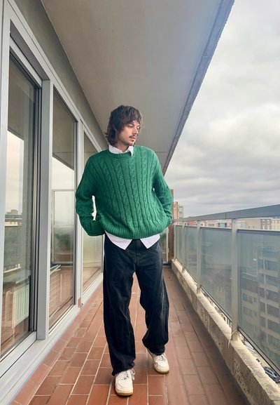 Suéter verde de punto con patrón de trenzas, superpuesto a una camisa blanca con cuello, combinado con pantalones negros de pierna ancha y zapatillas blancas.