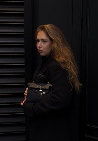 Sort læderhåndtaske med et struktureret design, guldfarvede metaldele og en toprem, båret af en person iført en sort frakke.