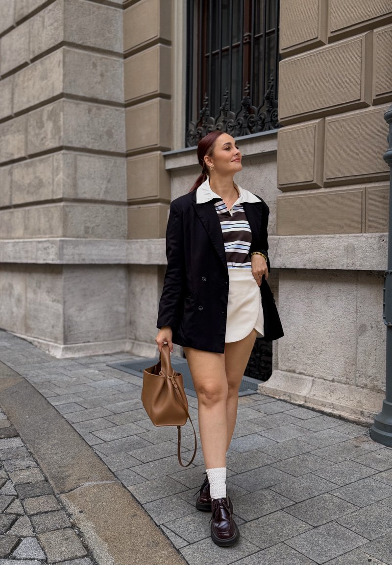 Blazer noir croisé sur une chemise rayée, jupe crème, sac à main en cuir marron et chaussures foncées avec des chaussettes blanches à la cheville. Cadre urbain.