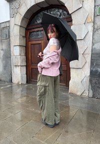 Νεαρή γυναίκα με παστέλ ροζ πουλόβερ και χακί παντελόνι cargo κρατάει μια μαύρη ομπρέλα σε υγρό λιθόστρωτο δρόμο μπροστά από μια ξύλινη καμάρα πόρτας.