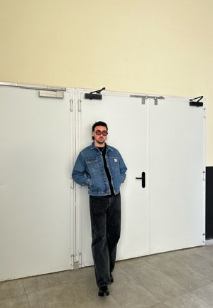 Chaqueta de mezclilla en un tono azul claro, superpuesta sobre una camiseta negra, combinada con pantalones de pierna ancha negros y zapatos negros, frente a puertas blancas.