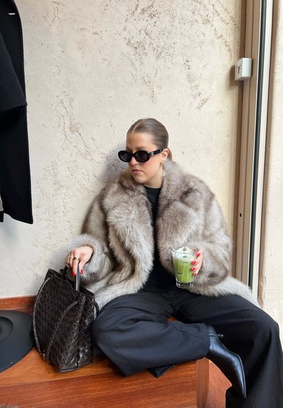 Abrigo de piel sintética en marrón claro con una textura suave. Bolso de mano negro trenzado, gafas de sol negras de gran tamaño, sosteniendo una bebida verde en un vaso.