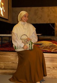 Femme portant un hijab beige et un collier de perles, assise avec un sac à main décoratif dans une pièce chaleureuse avec des coussins à motifs et une cheminée murale.
