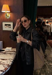 Must tumes nahkjakki, ümmargused prillid ja beež kott. Hoides selge kõrrega tassi. Marmorist leti ja puidust paneelide taust.